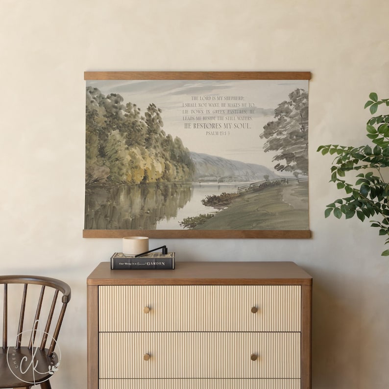 May include: A landscape print with a wooden frame, depicting a river scene with trees and a pathway. The text "The Lord is my shepherd" is printed on the artwork. The print hangs above a wooden dresser.