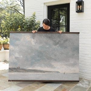 May include: A large rectangular framed artwork depicting a seascape with a cloudy sky. The painting features a muted color palette of grays, blues, and browns. The artwork is held by a person in front of a white brick wall.