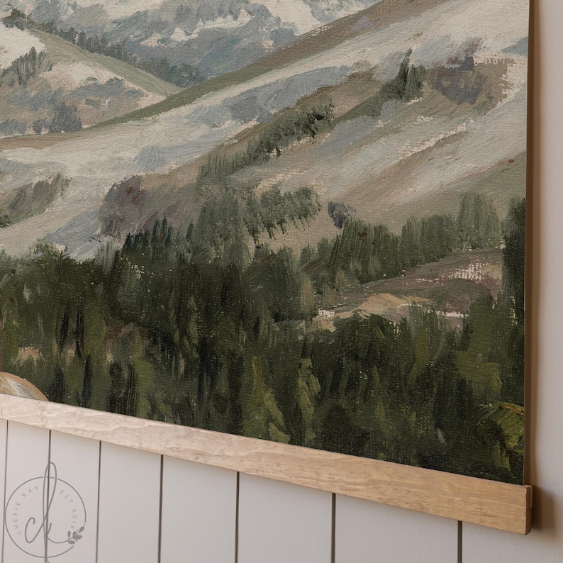 May include: An oil painting of a mountain landscape in muted tones of green, grey, and beige. The artwork depicts rolling hills and a forest, with a wooden frame at the bottom. The painting is mounted on a white wall.
