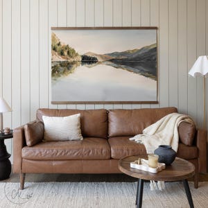 May include: A watercolor landscape painting of a lake and mountains hangs above a brown leather sofa. A white and beige striped pillow rests on the sofa. A round wooden coffee table holds a candle, a decorative tray, and a dark blue vase.