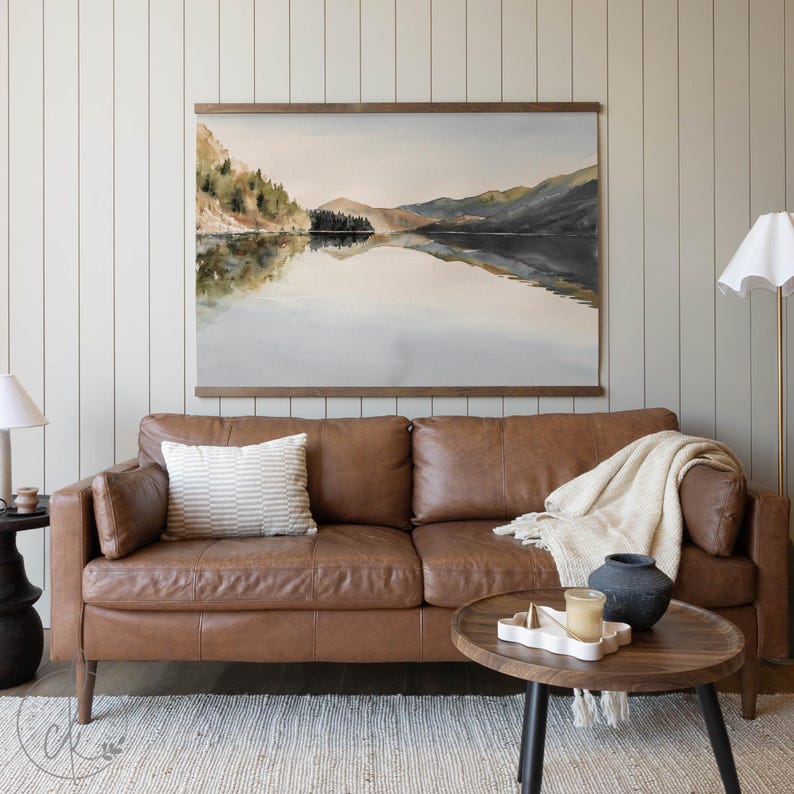 May include: A watercolor landscape painting of a lake and mountains hangs above a brown leather sofa. A white and beige striped pillow rests on the sofa. A round wooden coffee table holds a candle, a decorative tray, and a dark blue vase.