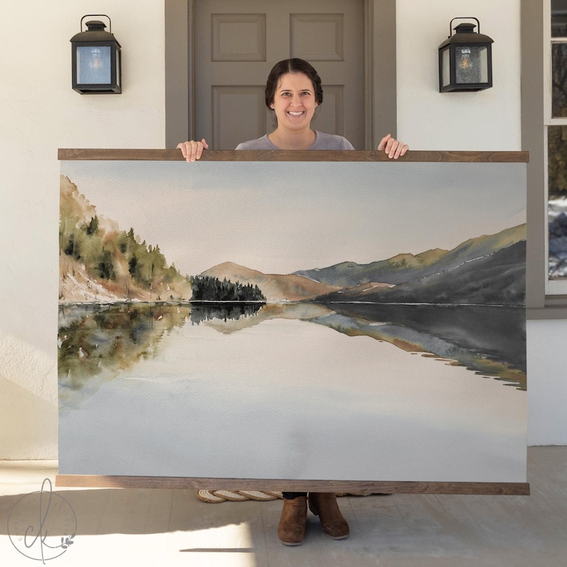 May include: A large watercolor landscape print featuring a serene lake scene with mountains and trees. The artwork is framed in a dark wood and is being held up by a person. The artwork is a landscape painting.