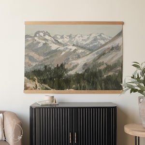 May include: A landscape painting depicting snow-covered mountains and a forest, presented in a wooden frame. The artwork uses a subdued colour palette of greys, greens, and browns, creating a tranquil, natural scene. The painting is mounted on a light-coloured wall.