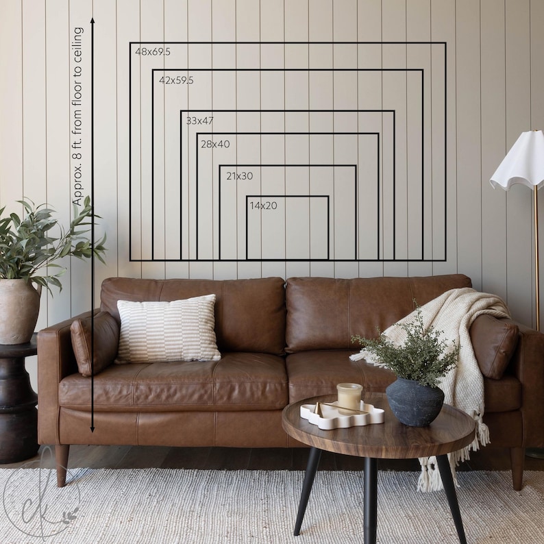 May include: A brown leather sofa with a striped pillow and a cream-colored throw blanket. A wooden coffee table with a decorative tray and a black vase with greenery. The wall displays framed rectangles with dimensions from 14x20 inches to 48x69.5 inches.