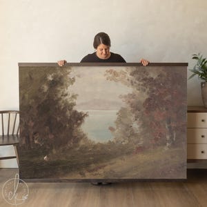 May include: Large framed landscape painting featuring a serene lake scene framed by trees. The artwork uses muted tones of green, brown, and blue, creating a vintage aesthetic. The frame is a dark wood.