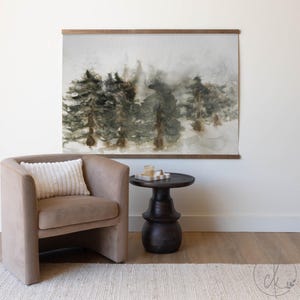 May include: Watercolor forest art print with a wooden frame. The artwork depicts a misty forest scene in shades of green and brown. The print is displayed above a tan armchair and a dark brown side table.
