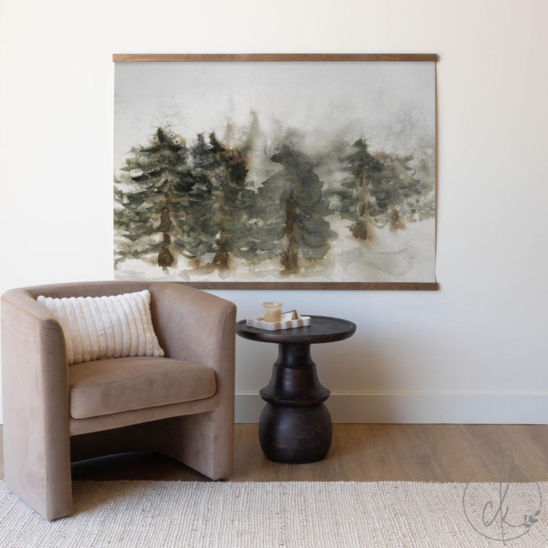 May include: Watercolor forest art print with a wooden frame. The artwork depicts a misty forest scene in shades of green and brown. The print is displayed above a tan armchair and a dark brown side table.
