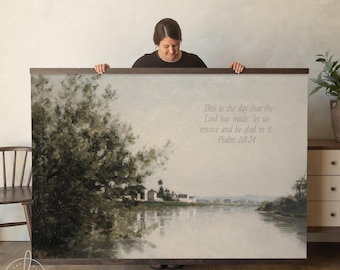 May include: A large rectangular landscape wall art print in a dark wood frame. The painting depicts a calm river scene with lush green trees on the left and distant buildings. The text reads: This is the day that the Lord has made; let us rejoice and be glad in it. Psalm 118:24.