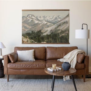 May include: A brown leather sofa with a striped cushion and a round wooden coffee table. A mountain landscape painting hangs above the sofa. A cream throw and a dark blue vase are on the table.