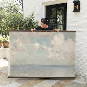 May include: Large rectangular artwork depicting a seascape with a cloudy sky. The painting features soft blues, grays, and whites, framed in a dark wood. The artwork is held by a person in front of a white brick wall.