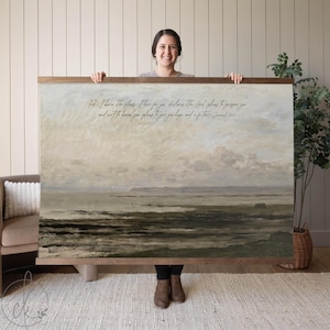 May include: A large framed landscape print with a muted color palette of beige, gray, and brown. The artwork features a coastal scene with text at the top. The frame is a natural wood tone. The print is held by a person.