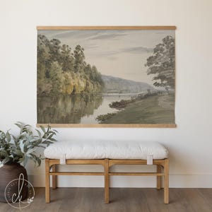May include: Watercolor landscape print of a river scene with trees and a mountain in the background. The artwork is displayed on a wooden frame. A light-colored bench with a white cushion sits below the artwork.