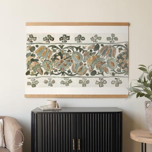 May include: A framed textile art piece featuring a floral design in muted tones of green, blue, and brown. The artwork is mounted on a light-colored wooden frame, creating a decorative wall hanging.