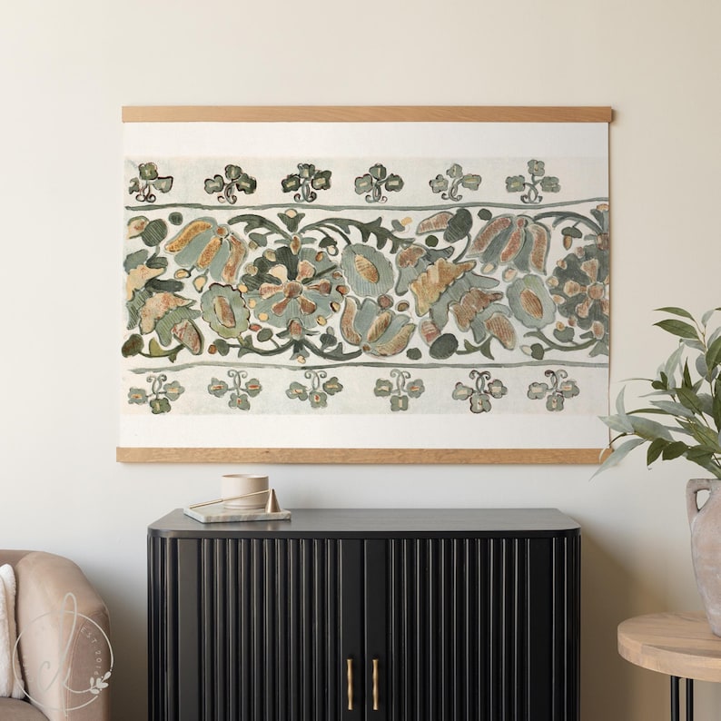 May include: A framed textile art piece featuring a floral design in muted tones of green, blue, and brown. The artwork is mounted on a light-colored wooden frame, creating a decorative wall hanging.