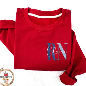 May include: A folded red sweatshirt with the embroidered letters "RN" in blue and white, with the name "Clara" in cursive script. The sweatshirt has a white collar and cuffed sleeves. The image includes an embroidery logo.