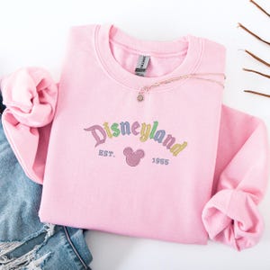 A pink sweatshirt with the text "Disneyland EST. 1955" printed on it, along with a Mickey Mouse logo, is displayed on a white background.