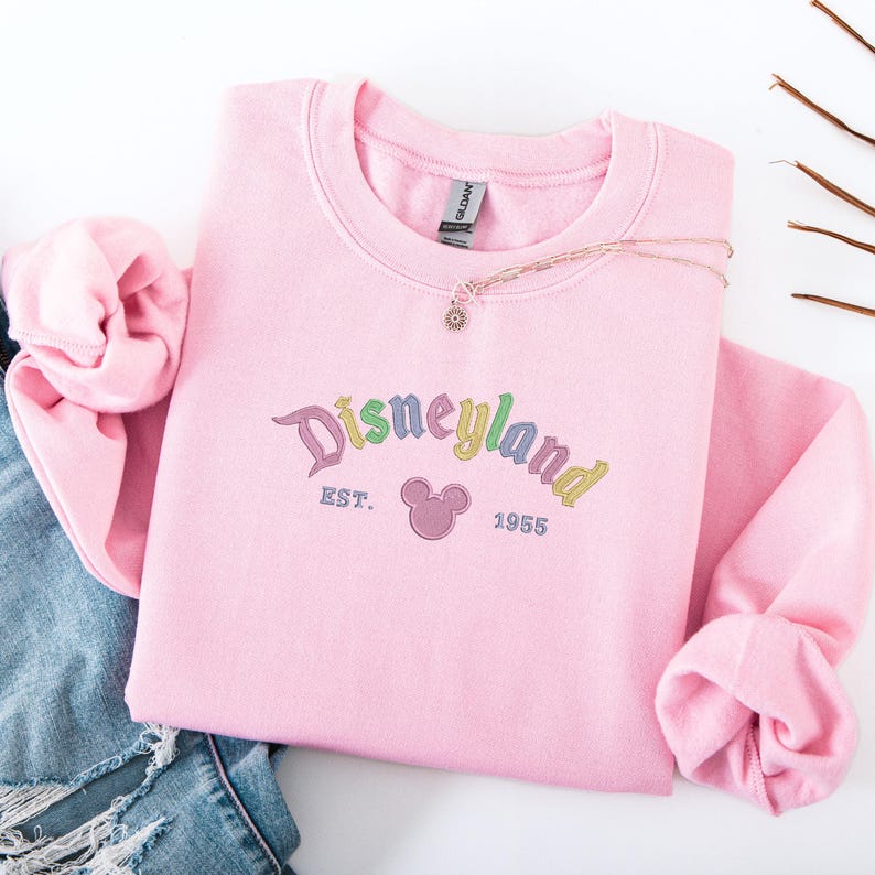 A pink sweatshirt with the text "Disneyland EST. 1955" printed on it, along with a Mickey Mouse logo, is displayed on a white background.