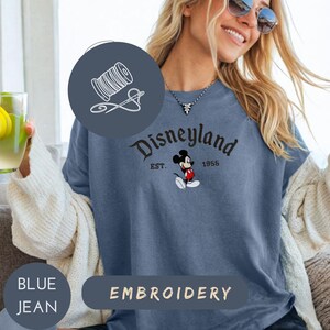 A woman wearing a blue t-shirt with a Mickey Mouse design and the text "Disneyland EST. 1955" is smiling and holding a drink.