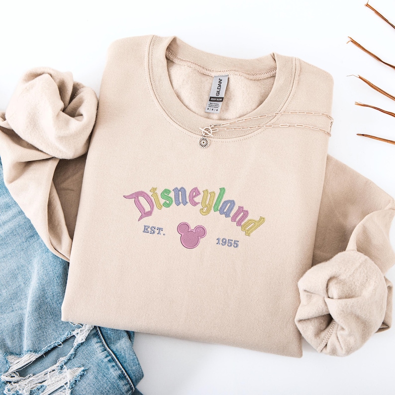 A beige sweatshirt with the text "Disneyland EST. 1955" printed on it, along with a Mickey Mouse logo, is displayed on a white background.