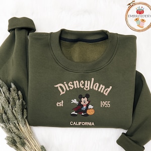 May include: An olive green sweatshirt featuring the word "Disneyland" in a script font, with "est 1955" and "CALIFORNIA" below. A Mickey Mouse graphic in a vampire costume is also present. The sweatshirt has a crew neck and long sleeves.