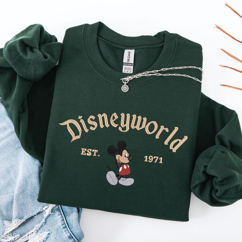 A green sweatshirt with the text "Disneyworld EST. 1971" printed on it, along with a Mickey Mouse graphic. The sweatshirt is placed on a white background, and there is a pair of denim jeans visible in the background.