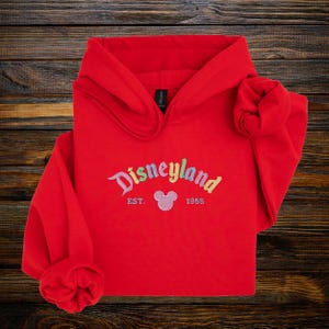 A red hoodie with the text "Disneyland EST. 1955" printed on it, along with a Mickey Mouse logo, is displayed on a wooden surface.
