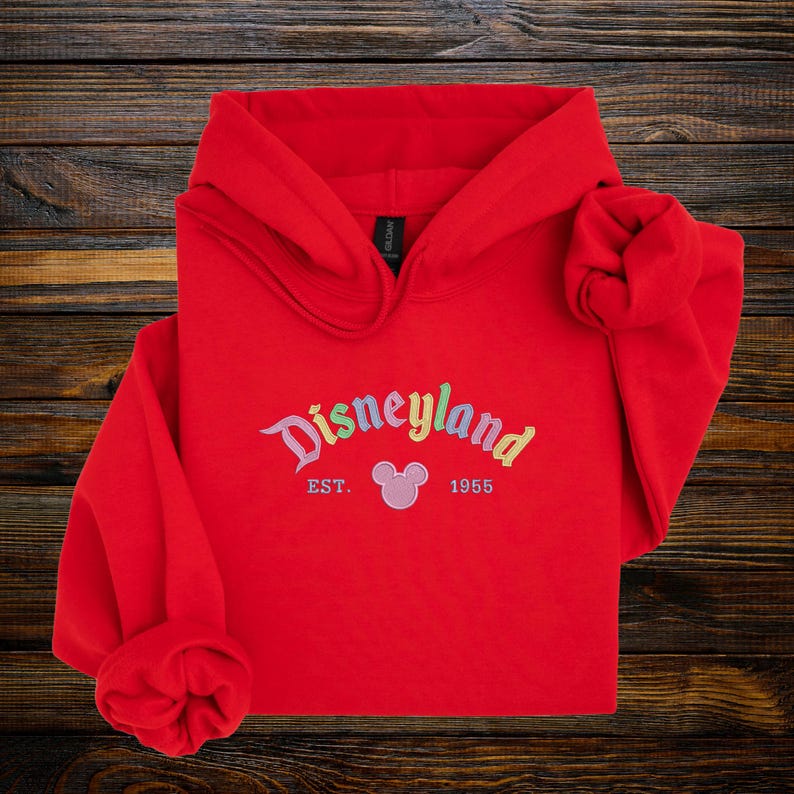 A red hoodie with the text "Disneyland EST. 1955" printed on it, along with a Mickey Mouse logo, is displayed on a wooden surface.