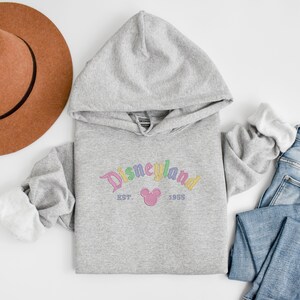 A gray hoodie with the text "Disneyland EST. 1955" printed on it, along with a brown hat and a pair of blue jeans, all laid out on a white background.