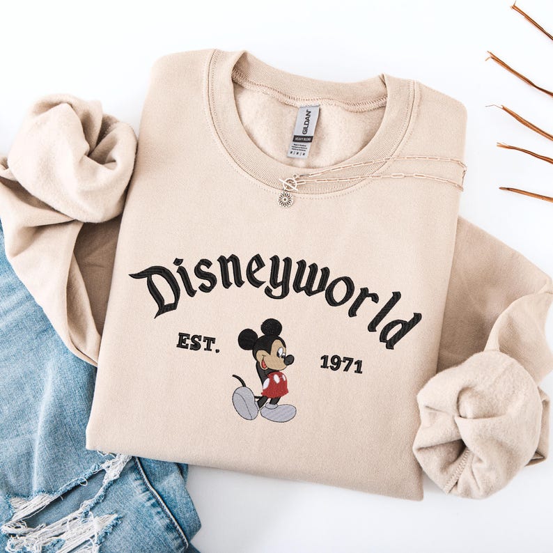 A beige sweater with the text "Disneyworld EST. 1971" printed on it, along with a Mickey Mouse graphic, is laid out on a white background.