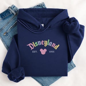 A navy blue hoodie with the word "Disneyland" printed on it, along with the year "1955" and a Mickey Mouse logo, is displayed on a white background.