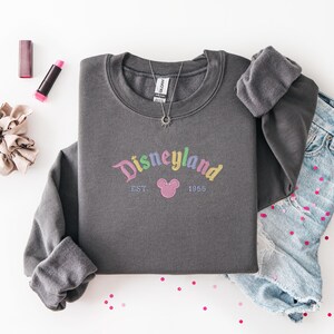 a gray sweatshirt with the text "Disneyland EST 1965" printed on it, along with a pair of denim jeans and some makeup items, such as lipstick and a hair tie, arranged on a white background.