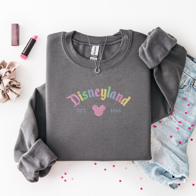a gray sweatshirt with the text "Disneyland EST 1965" printed on it, along with a pair of denim jeans and some makeup items, such as lipstick and a hair tie, arranged on a white background.
