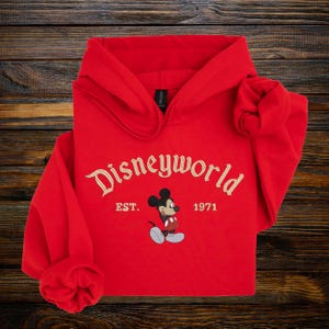 A red hoodie with the text "Disneyworld EST. 1971" and a Mickey Mouse graphic printed on it, laid out on a wooden surface.