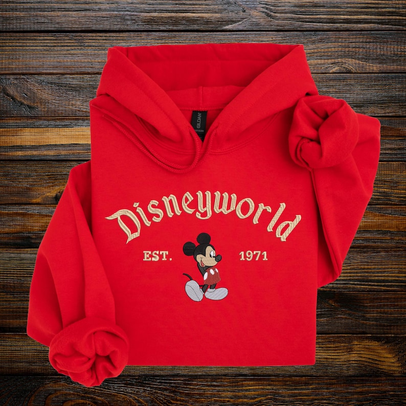 A red hoodie with the text "Disneyworld EST. 1971" and a Mickey Mouse graphic printed on it, laid out on a wooden surface.