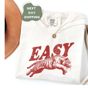 May include: Cream-colored t-shirt with the word "EASY" in red, above a red tiger graphic. The shirt has a crew neck and short sleeves. A gold necklace with a pendant is draped on the shirt.