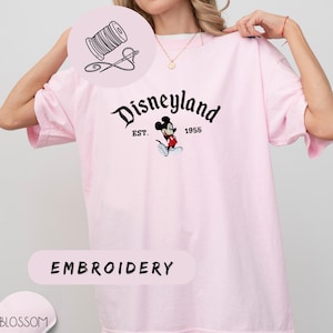 A woman wearing a pink t-shirt with the Disneyland logo and text, posing with her hand on her shoulder.