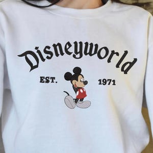 A person wearing a white sweatshirt with the text "Disneyworld EST. 1971" and a Mickey Mouse graphic printed on it.