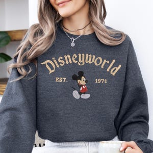 A woman wearing a gray sweatshirt with the text "Disneyworld EST. 1971" and a Mickey Mouse graphic printed on it.
