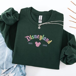 A green sweatshirt with the text "Disneyland EST. 1955" printed on it, along with a Mickey Mouse logo, is displayed on a white background.