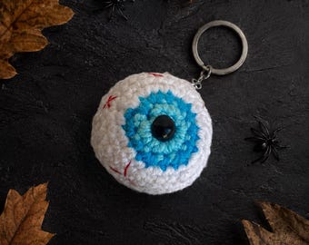 May include: A round, crocheted eyeball keychain featuring a white sclera with red embroidered veins, a bright blue iris, and a central black pupil. The plush eyeball is attached to a circular silver-toned metal split ring for keys.