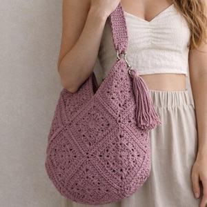 Könnte beinhalten: Eine staubrosa gehäkelte Schultertasche mit geometrischem Muster. Die Tasche hat einen passenden Riemen und eine dekorative Quaste. Die Tasche wird von einer Person gehalten. Die Tasche ist aus weichem, strukturiertem Garn gefertigt.