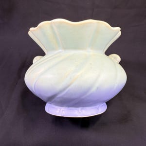 May include: A ceramic vase with a seashell design. The vase has a light blue and lavender color scheme with a ruffled rim and two small handles. The base of the vase has a decorative trim.