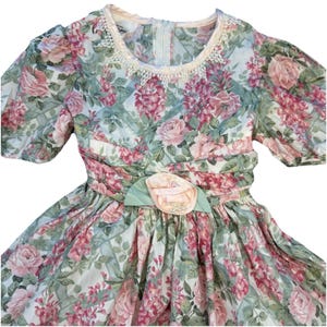 May include: A short-sleeved dress featuring a floral pattern with pink roses and green leaves on a white background. The dress has a round neckline with white lace trim and a gathered waist accented by a pale pink fabric flower and green leaves.