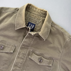 May include: A close-up shot of a khaki-coloured GAP jacket. The jacket features a collared neckline, two front pockets, and button fastenings. The GAP logo is visible on a label near the collar. The fabric appears to be a durable cotton or cotton blend.