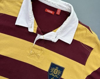 Puede incluir: Una camiseta de rugby a rayas burdeos y doradas con cuello blanco. La camiseta presenta un pequeño escudo bordado en el pecho y la marca "IZOD" en una etiqueta cerca del cuello.