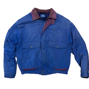 May include: A blue jacket with a burgundy collar and trim. The jacket has two front pockets with button closures and a zipper closure. The jacket has a quilted lining and is made of a durable material.