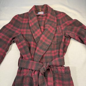 May include: A long-sleeved robe featuring a dark red and black plaid pattern. The garment includes a wide shawl collar, a matching fabric tie belt secured at the waist, and a single patch pocket on the upper left chest area of the front panel.