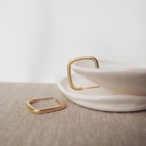 Op de afbeelding: Twee goudkleurige vierkante oorringen. Eén oorring ligt op een wit keramisch bord, de andere op een beige stoffen oppervlak. De oorbellen hebben een minimalistisch, geometrisch ontwerp.