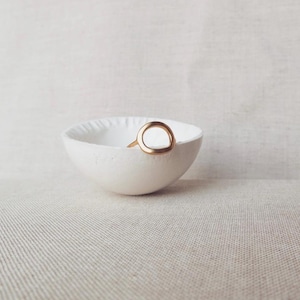 Peut inclure: Un petit bol en céramique blanche à surface texturée. Une poignée en forme d'anneau de couleur dorée est fixée sur le côté. Le bol est posé sur un fond neutre, créant une esthétique minimaliste. Idéal pour contenir de petits objets.