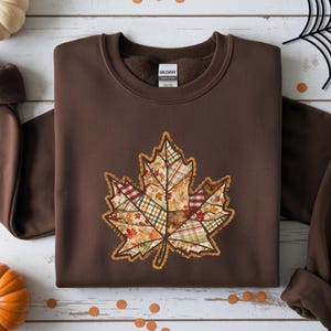 May include: A brown sweatshirt featuring a large, patchwork-style maple leaf design in autumn colors. The leaf has a variety of patterns, including plaid and floral prints. The sweatshirt is laid flat on a white wooden surface, with pumpkins and fall decorations.